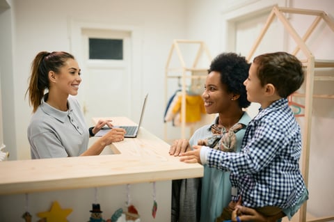 Happy kindergarten teacher communicating with African American mother and her son at reception desk.